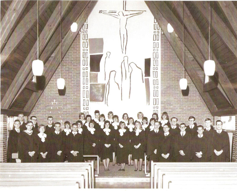 A choir, wearing robes, posed at the front of a church with a beautiful, modern mural.